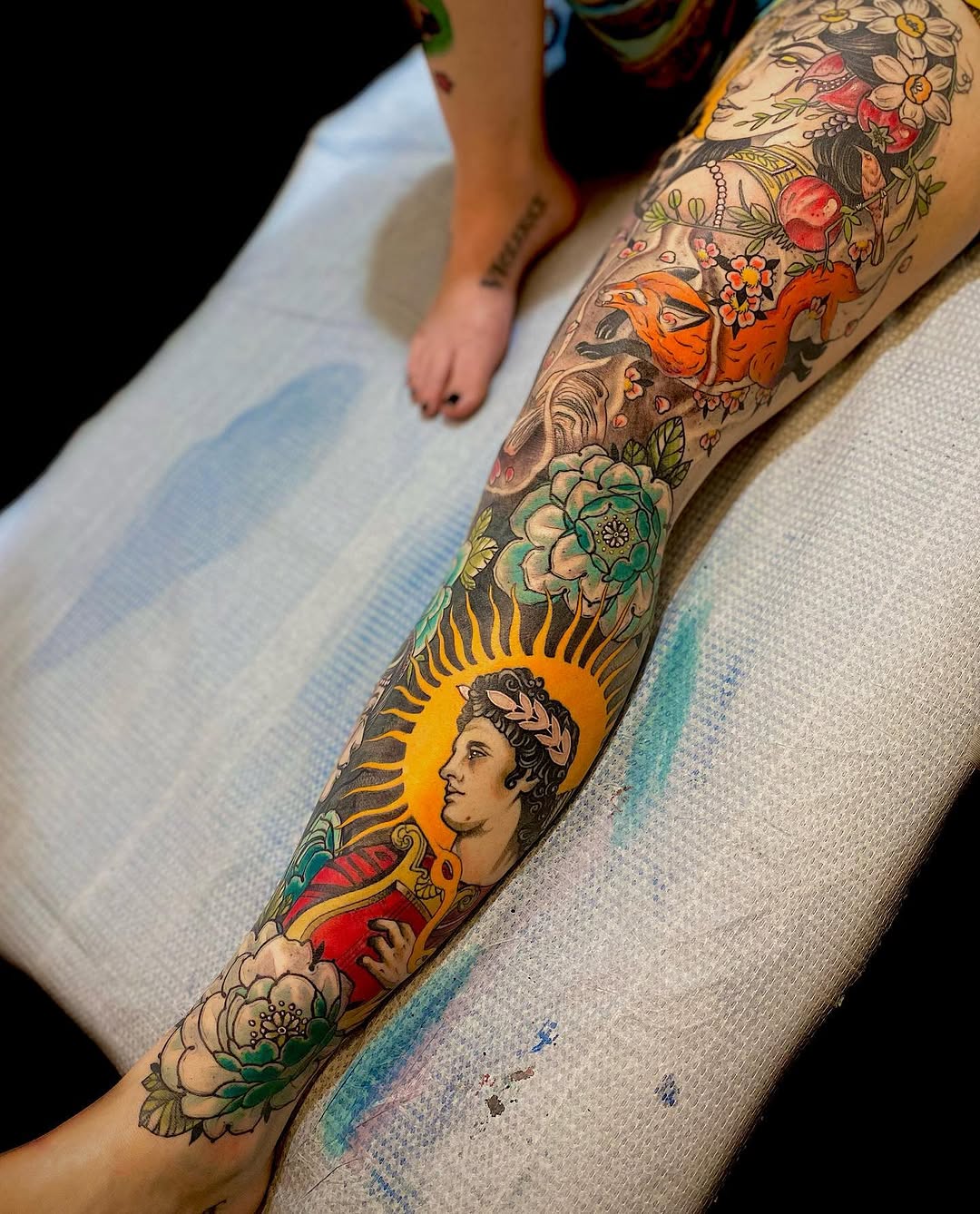 Full-leg sleeve tattoo featuring a classical male bust with halo, flowers, an orange bird, and sun motif in vibrant colors on pale skin.