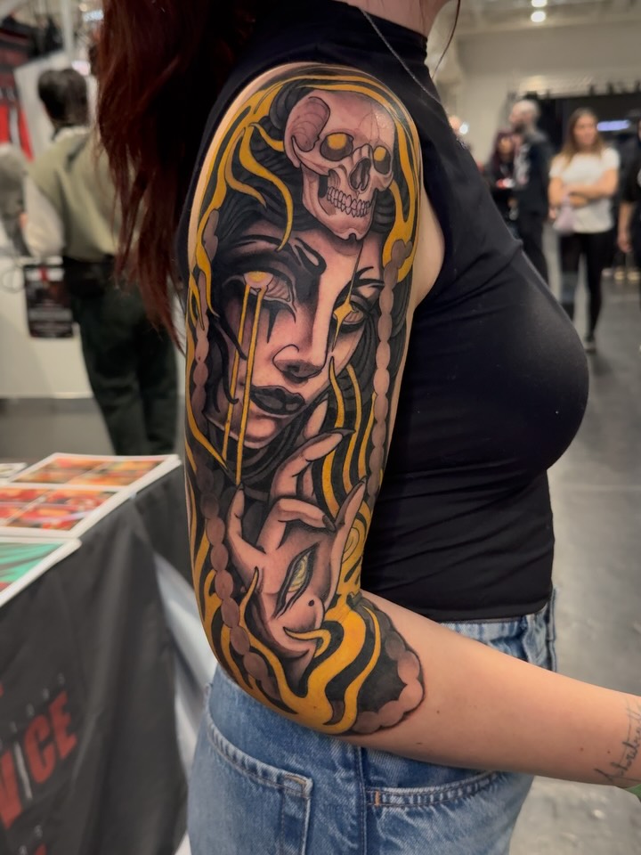 Large sleeve tattoo featuring a skull at the top, female faces with gold and black flowing elements extending down the arm, displayed on a person wearing a black shirt.