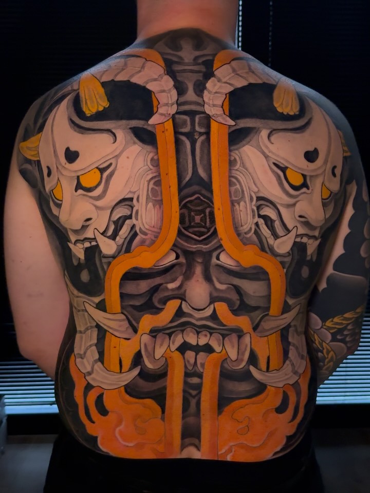 Full back tattoo featuring symmetrical demon masks with orange accents, interconnected by orange ribbons and geometric patterns down the spine.