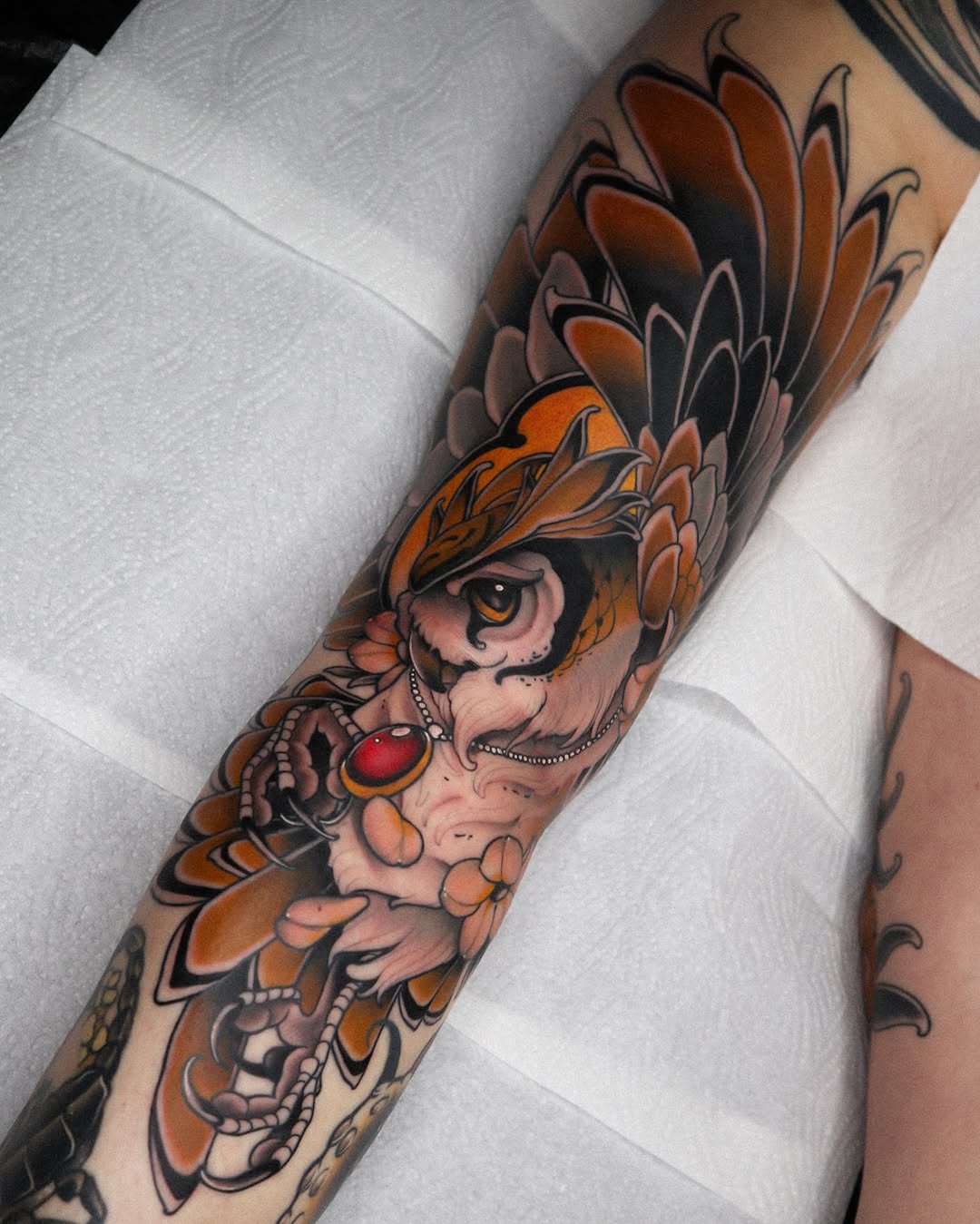 Colorful arm tattoo featuring an owl with orange and brown feathers, holding a red jeweled orb, surrounded by decorative floral elements.