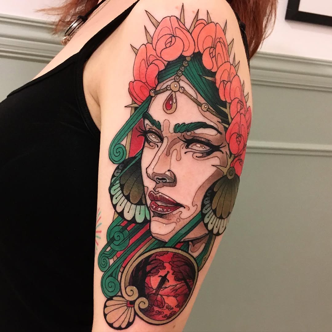 Colorful shoulder tattoo featuring a face with green feathered headdress, coral roses, closed eyes with tears, and a red gemstone pendant.