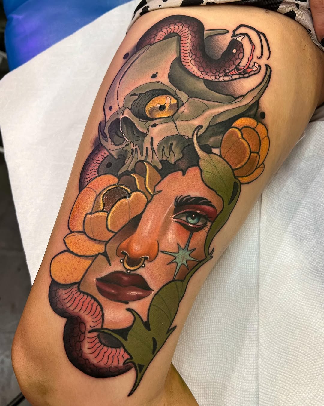 Colorful shoulder tattoo featuring a woman's face with skull overlay, serpents, orange flowers, and green leaves in warm and cool tones.