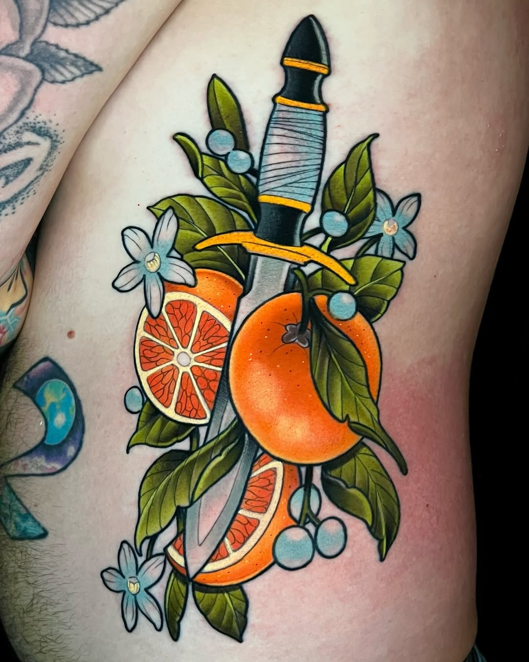Colorful tattoo on upper arm featuring oranges and grapefruit slices, green leaves, small blue flowers, and a dagger with wrapped handle and gold accents.