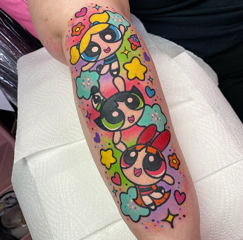 Three cartoon superhero girls with black masks and blonde/dark hair surrounded by colorful stars, hearts, and flowers on a calf tattoo in bright pink, yellow, green, and blue.