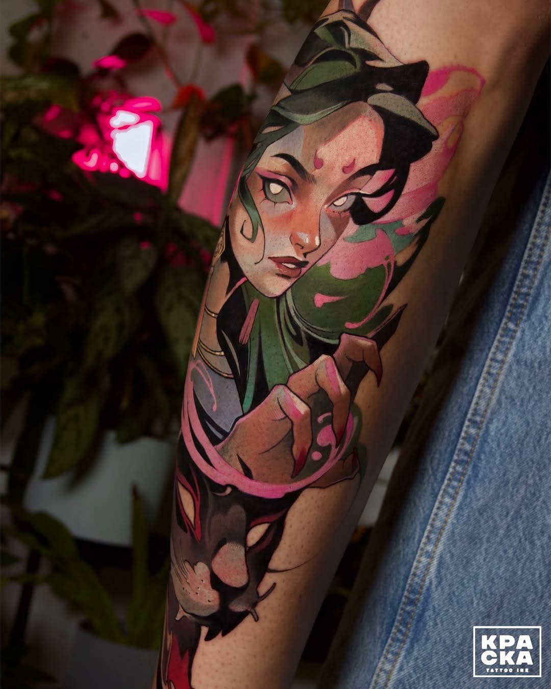 Colorful tattoo on an upper arm featuring a stylized female face with dramatic eyes, wearing a green headwrap, surrounded by pink floral elements and warm skin tones.