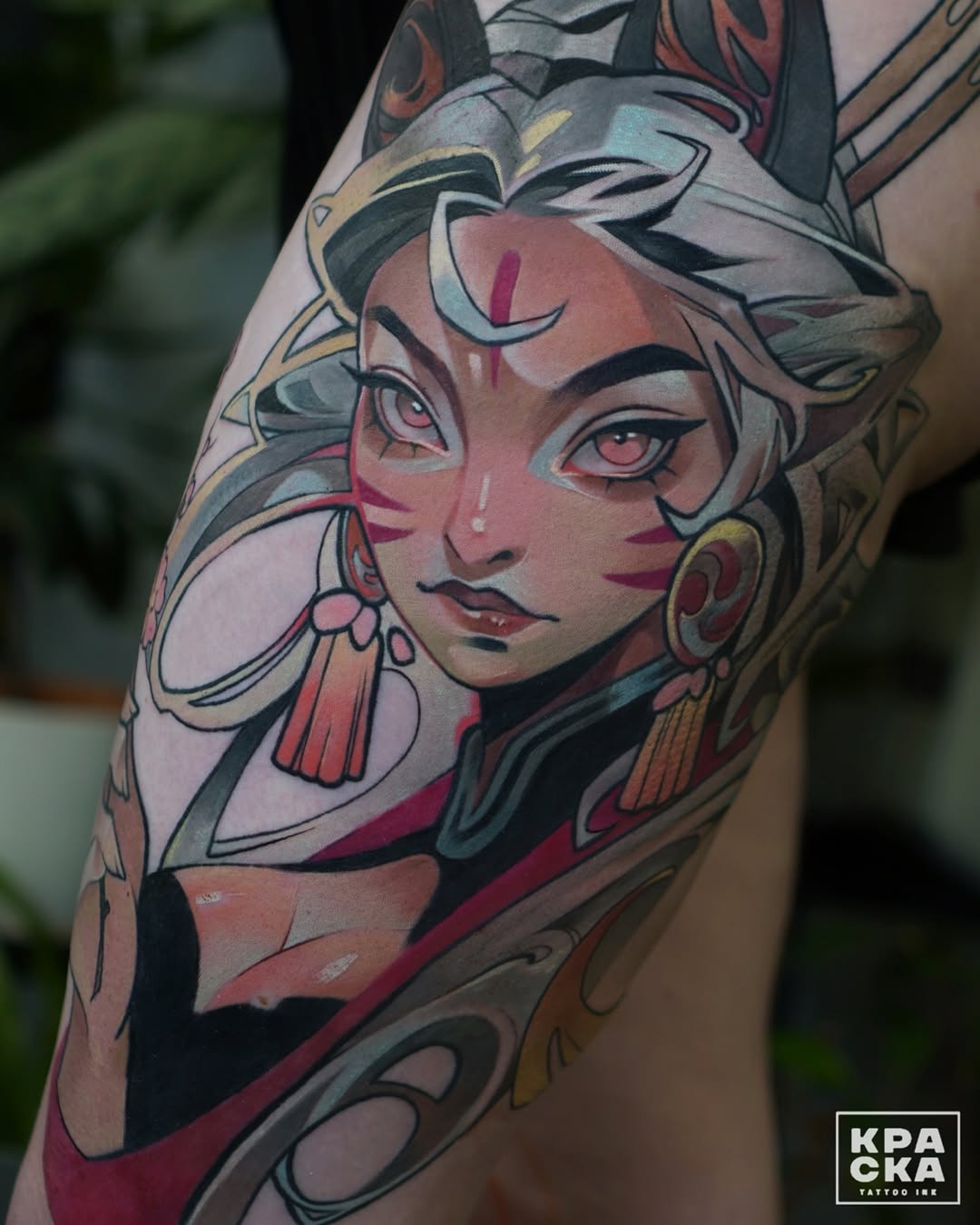Colorful shoulder tattoo featuring a stylized female face with pink and gray tones, decorative earrings, and ornamental elements framing the portrait.