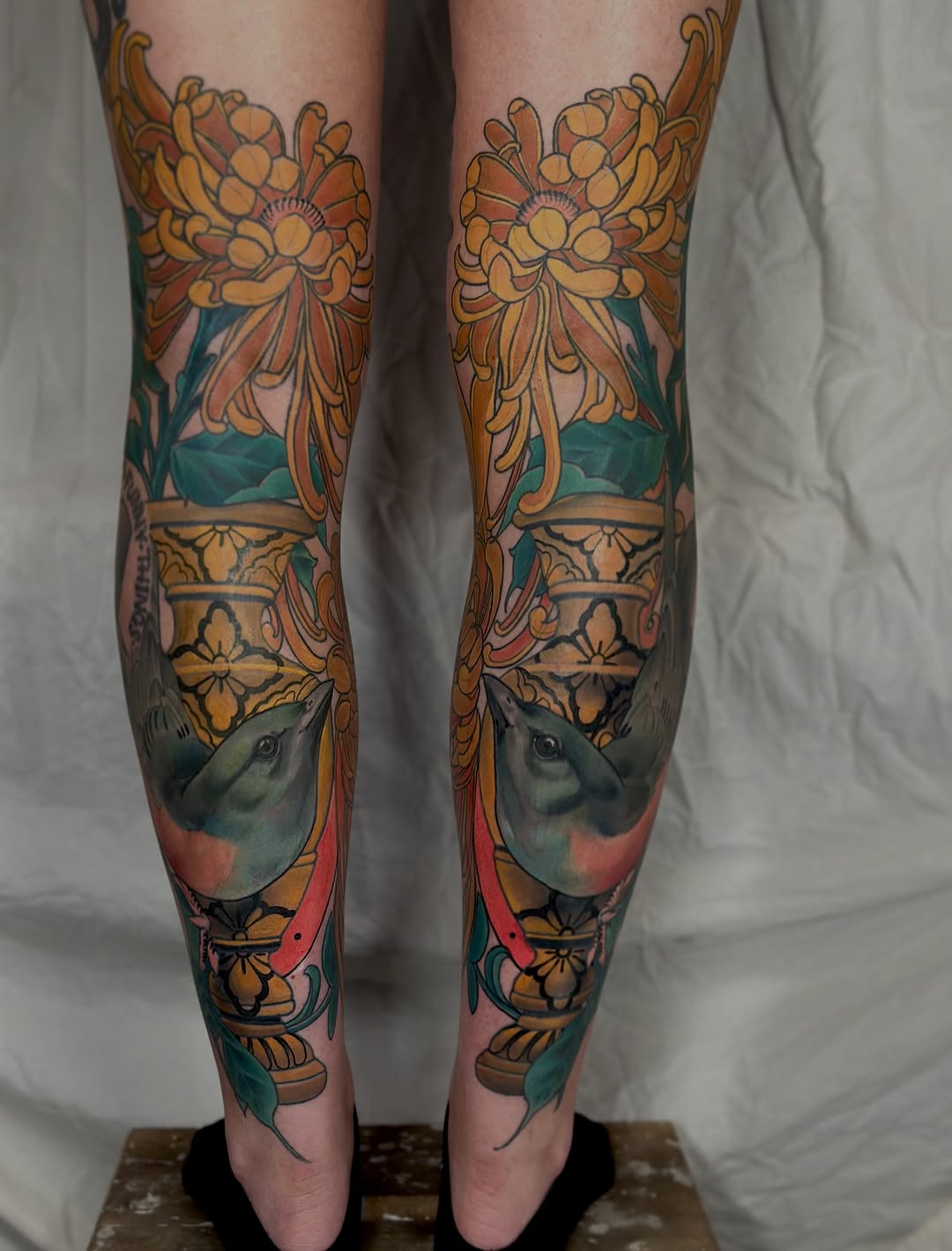 Matching leg tattoos featuring ornate floral urns with bird portraits, rendered in gold, teal, and coral colors on both legs.