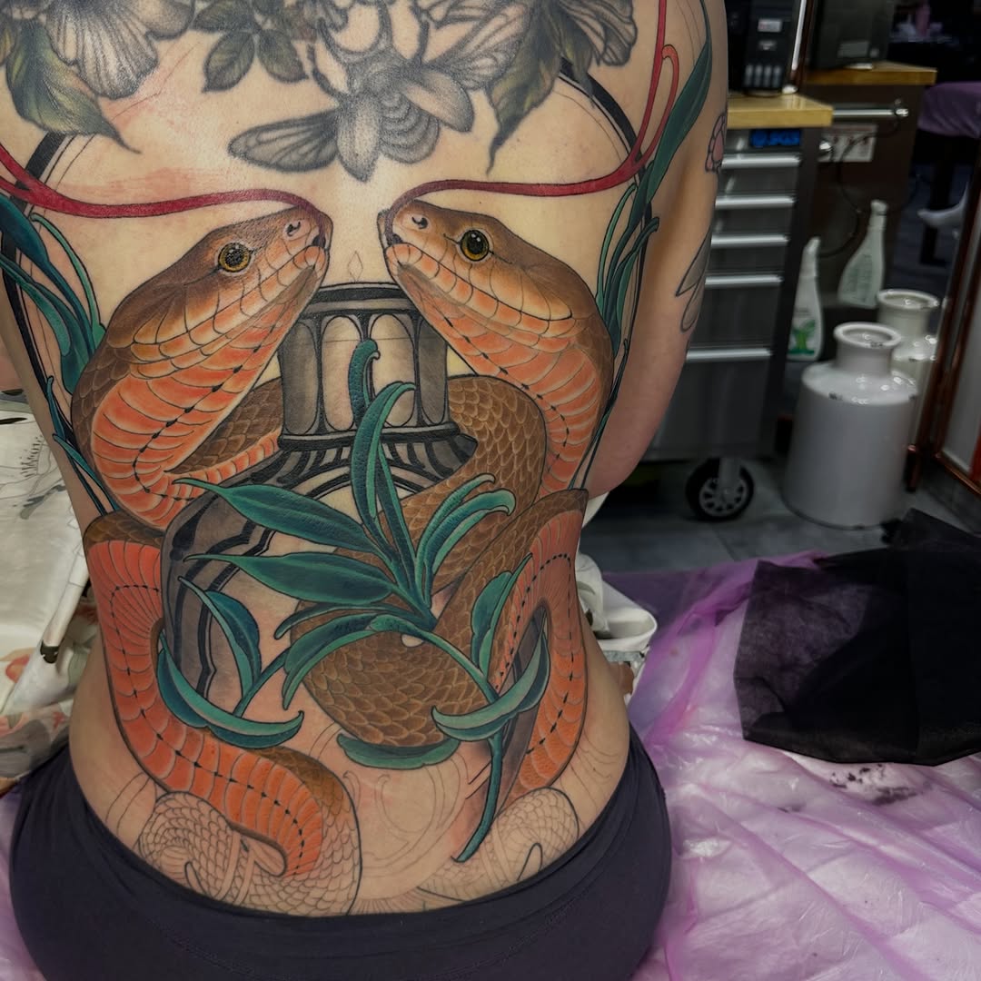 Two orange snakes with green eyes coiled around green foliage and a geometric symbol on the upper back and torso, with floral elements above.