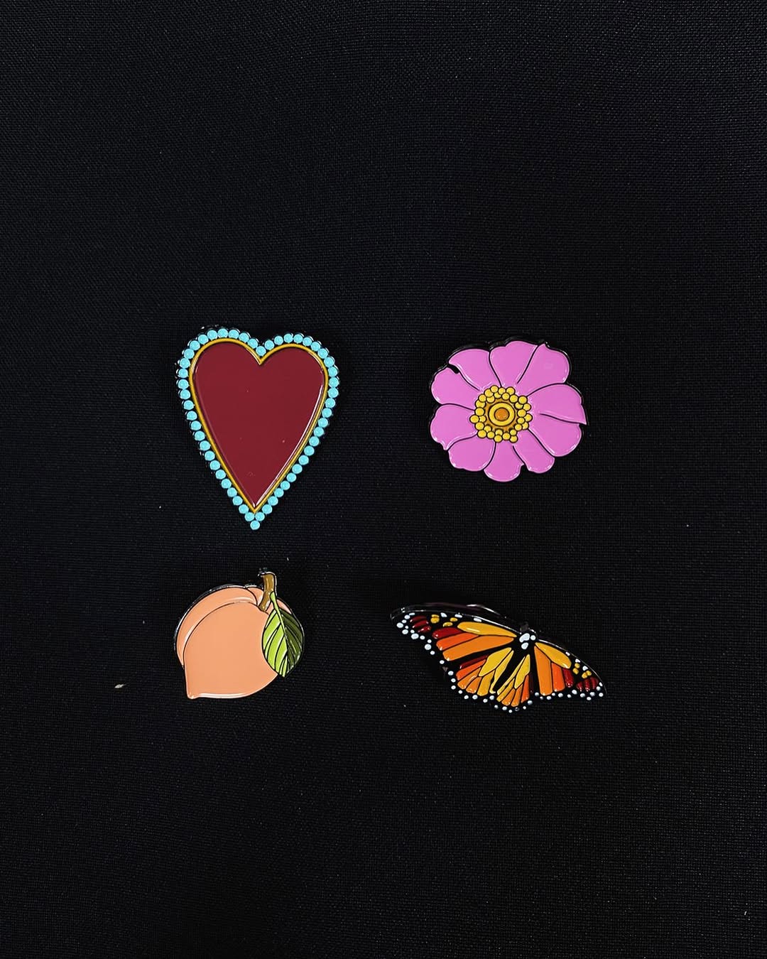 Four colorful tattoo designs on black fabric: a red heart with turquoise jeweled border, pink flower with yellow center, peach fruit with leaf, and orange monarch butterfly.