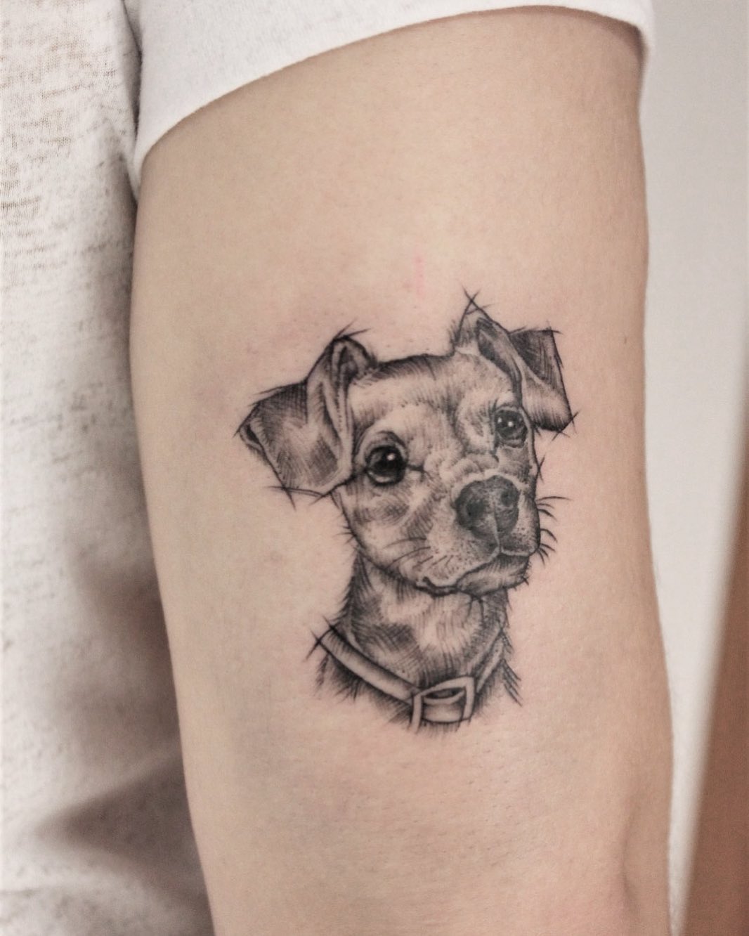 Small dog head portrait tattoo on upper arm in black ink, depicting a tilted dog wearing a collar, rendered with detailed linework.