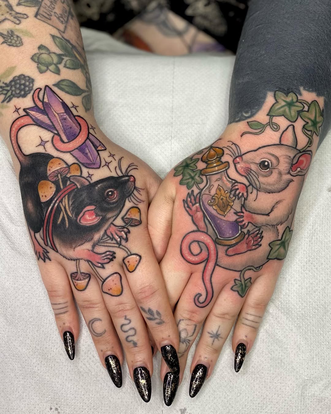 Two hands displaying matching complementary tattoos of rats holding food and wearing clothing, surrounded by vines and mushrooms in purple, orange, and green ink.