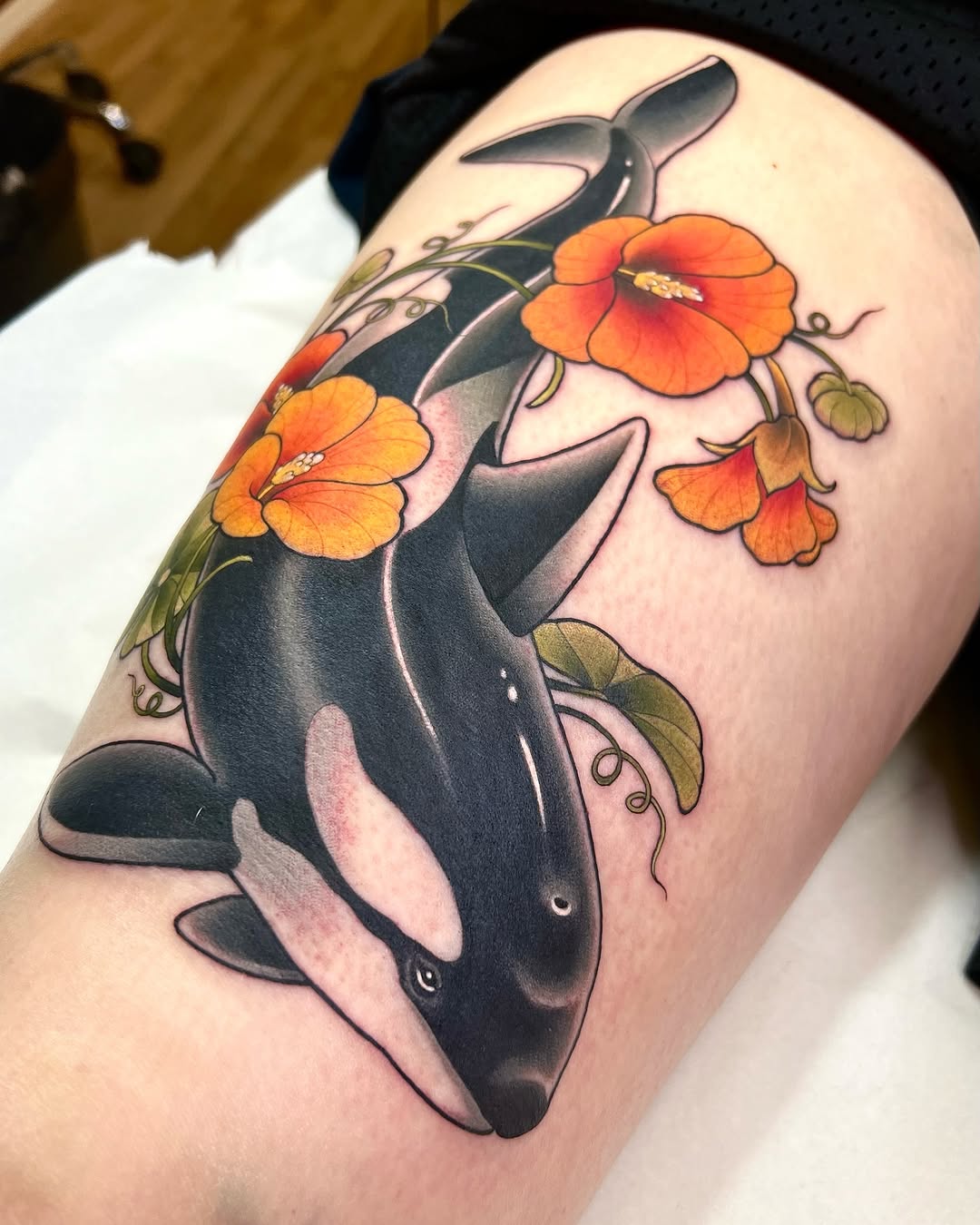 Orca whale with orange and yellow flowers and green leaves wrapped around its body, tattooed on an upper arm.