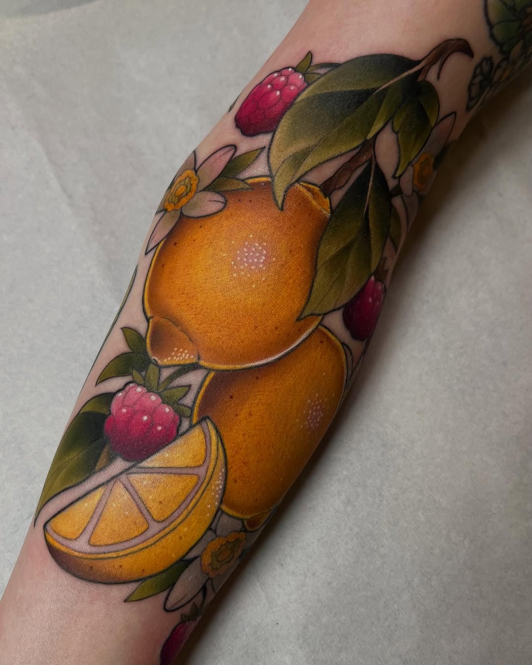 Colorful arm tattoo featuring oranges, raspberries, lemon slices, and green leaves with small flowers arranged vertically on the forearm.