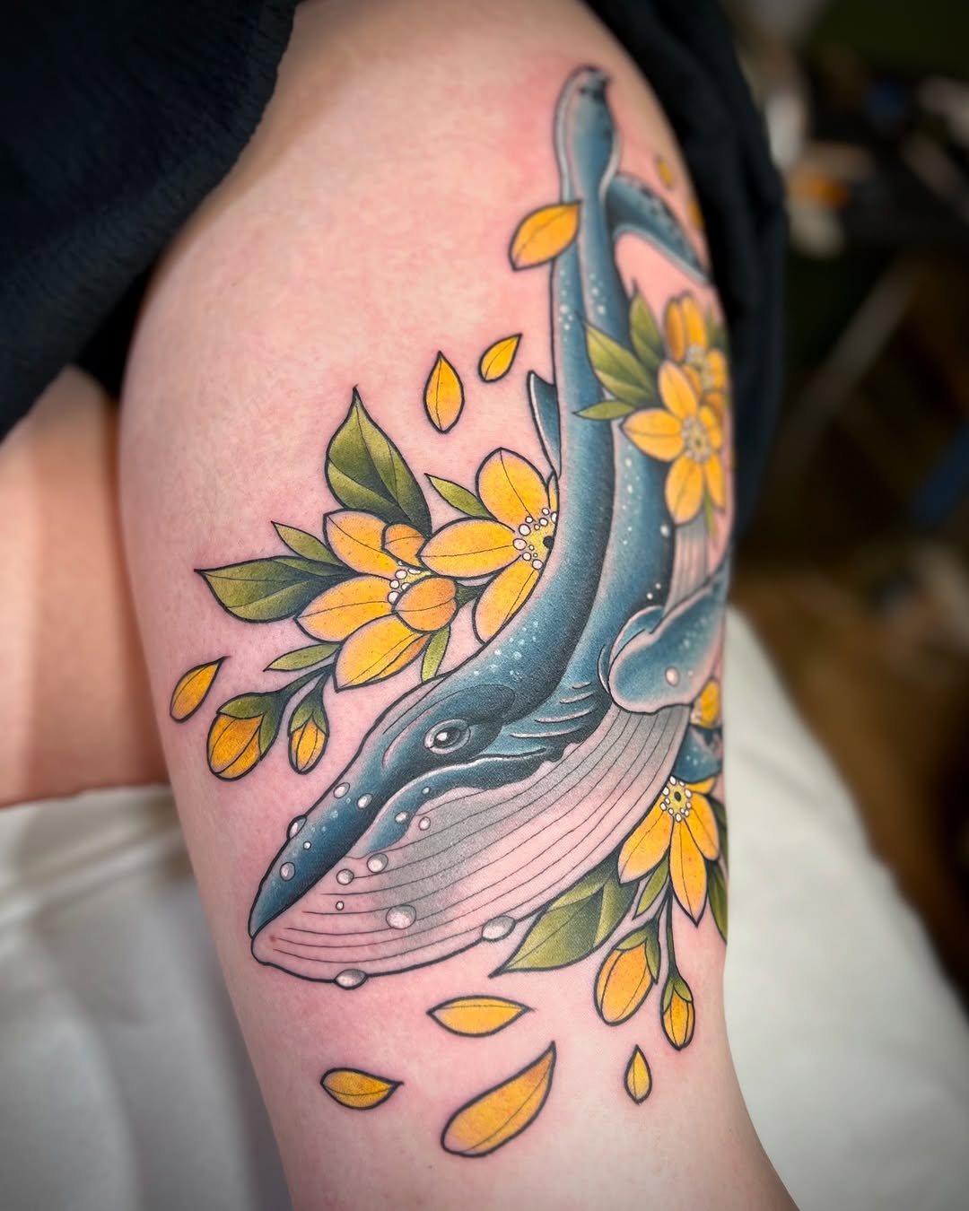 Whale tattoo with yellow flowers and falling petals on upper arm, featuring blue and gray whale with green leaves and white details.