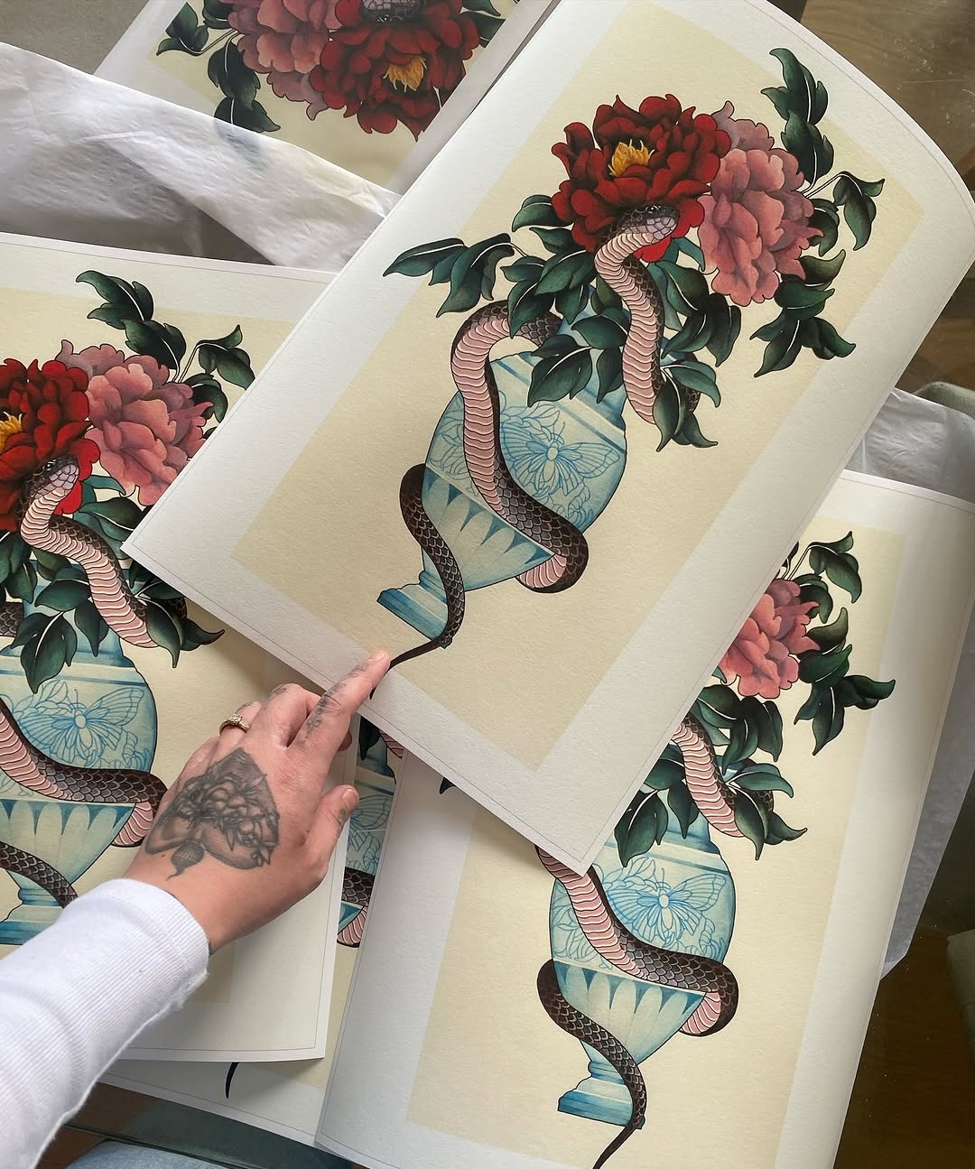 Printed portfolio samples displaying snakes coiled around decorative blue vessels with red and pink peonies, green leaves, and yellow flower centers on cream paper.