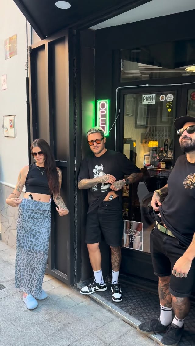 Three people with visible arm and leg tattoos stand in the doorway of a tattoo studio with a green neon sign.