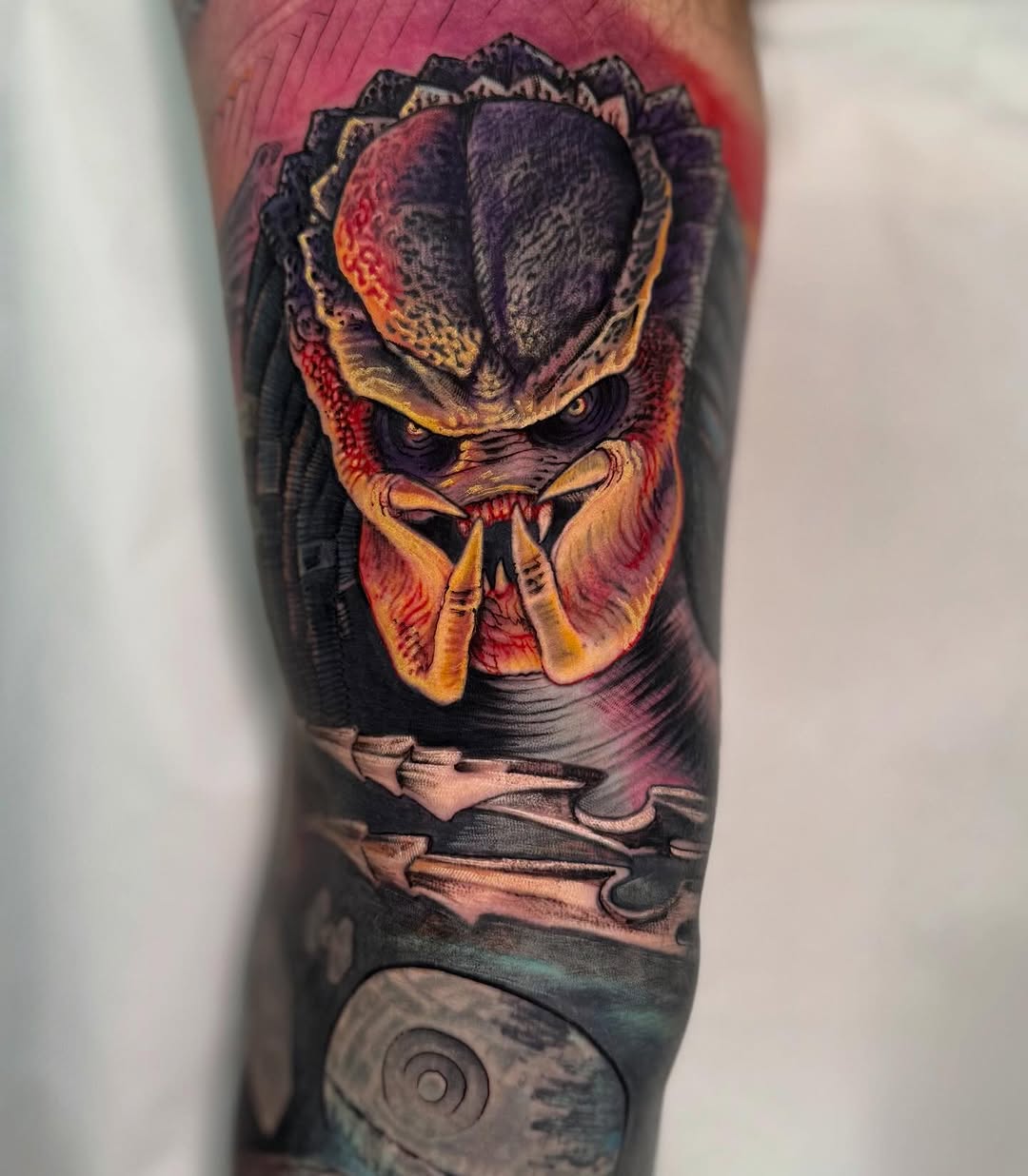 Colorful Predator creature head tattoo on leg featuring red, gold, and black tones with textured details and spiral design at bottom.
