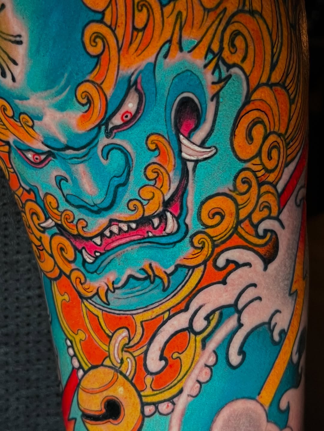 Colorful tattoo on leg featuring a turquoise demon mask with pink mouth, white teeth, and orange decorative scrollwork against skin.