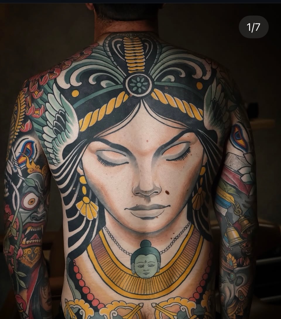 Large-scale female face tattoo on back with ornate headdress, wings, and jewelry in gold, teal, black, and cream colors.