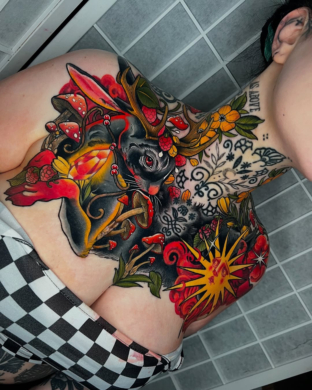 Colorful shoulder tattoo featuring a black cat surrounded by red roses, yellow flowers, mushrooms, and ornamental swirls in red, yellow, orange, and green.