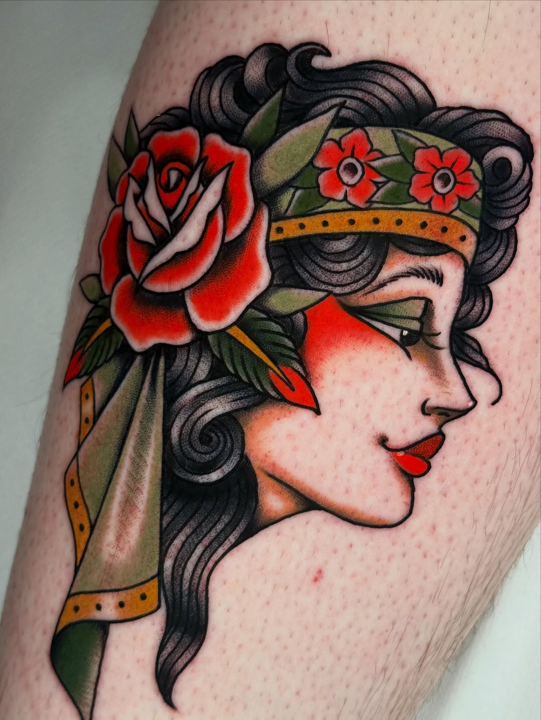 Blindfolded female profile head with black wavy hair, red rose, red flowers on golden band, and green leaf accents tattooed on upper arm.