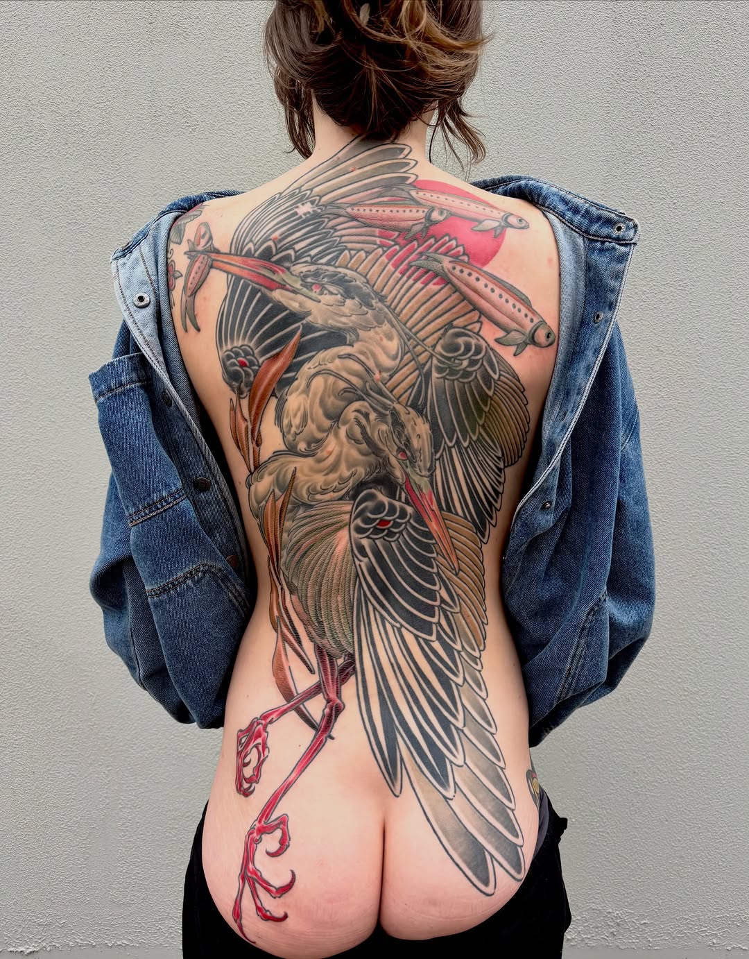 Large-scale back tattoo featuring winged figures, flying fish, and ornamental elements in red, black, and grey tones spanning the spine and shoulders.