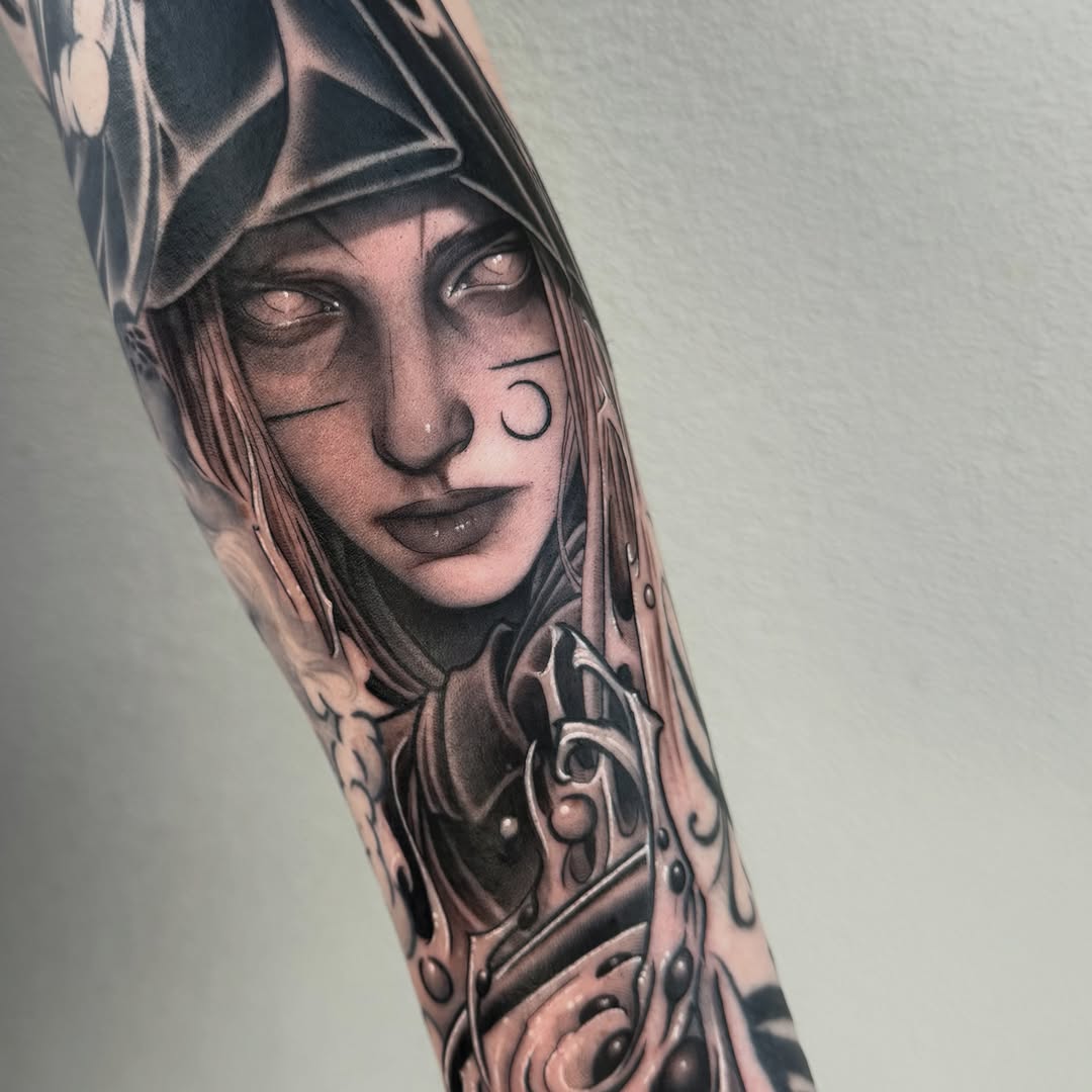 Detailed portrait tattoo on forearm featuring a woman's face with geometric patterns above, ornamental designs below, rendered in black and grey ink tones.