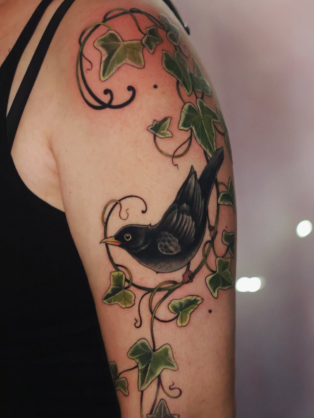 A black bird perched among green ivy vines with curling tendrils on the upper arm, against peachy skin tones.