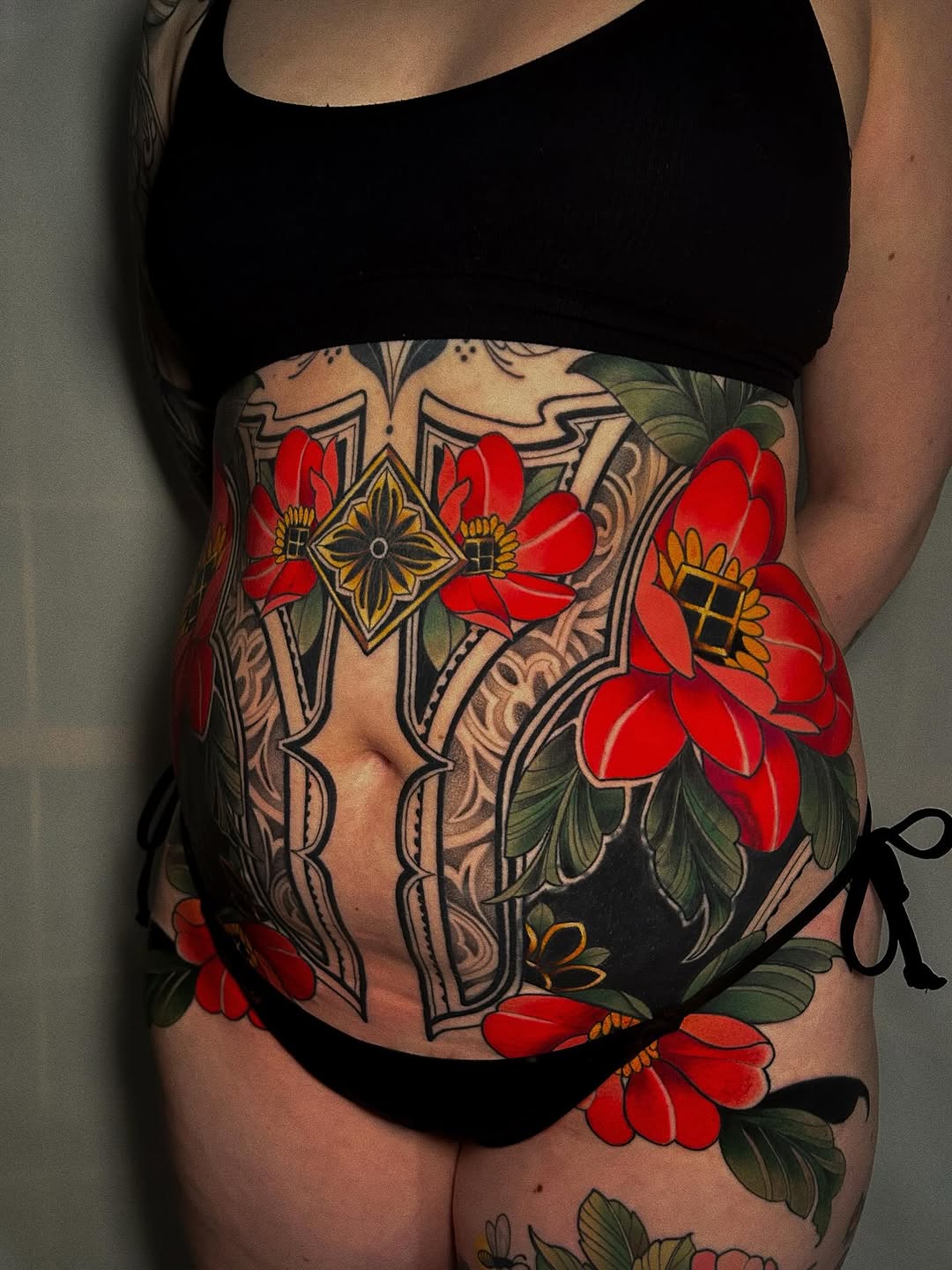 Colorful torso tattoo featuring red flowers with yellow centers, ornate geometric patterns, and decorative line work in gold and black tones.