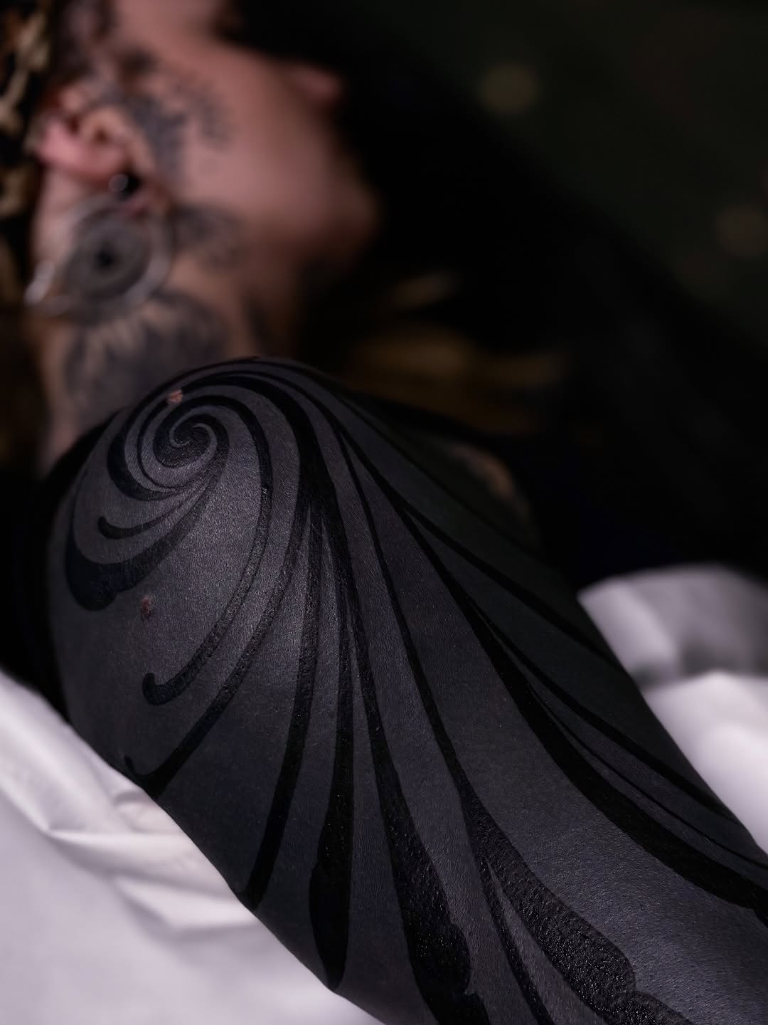 Spiral and curved line tattoo in black ink on a person's shoulder, with blurred background showing additional body art.