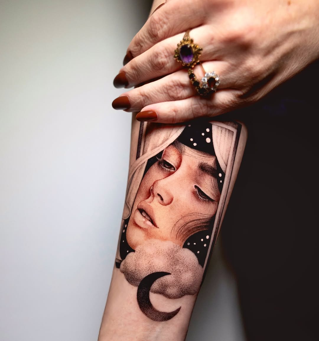 Portrait tattoo on forearm featuring a contemplative face with closed eyes, crescent moon, stars, and decorative elements in brown and black ink.