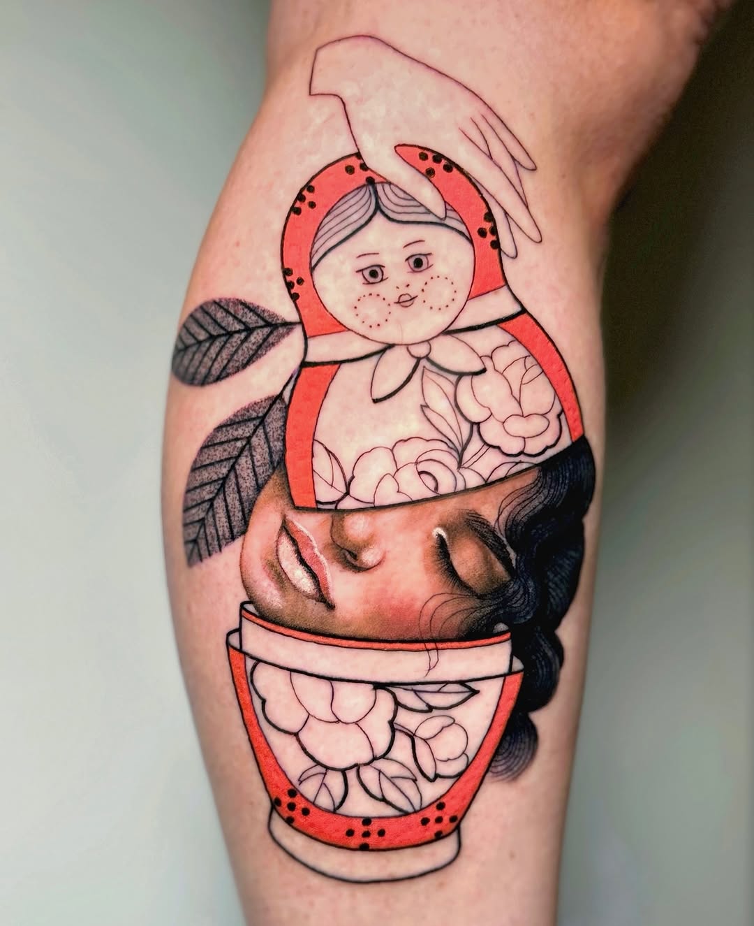 Colorful leg tattoo featuring a nesting doll design with a red-clothed figure on top, a face in the middle, and floral patterns, accented with black leaf details.
