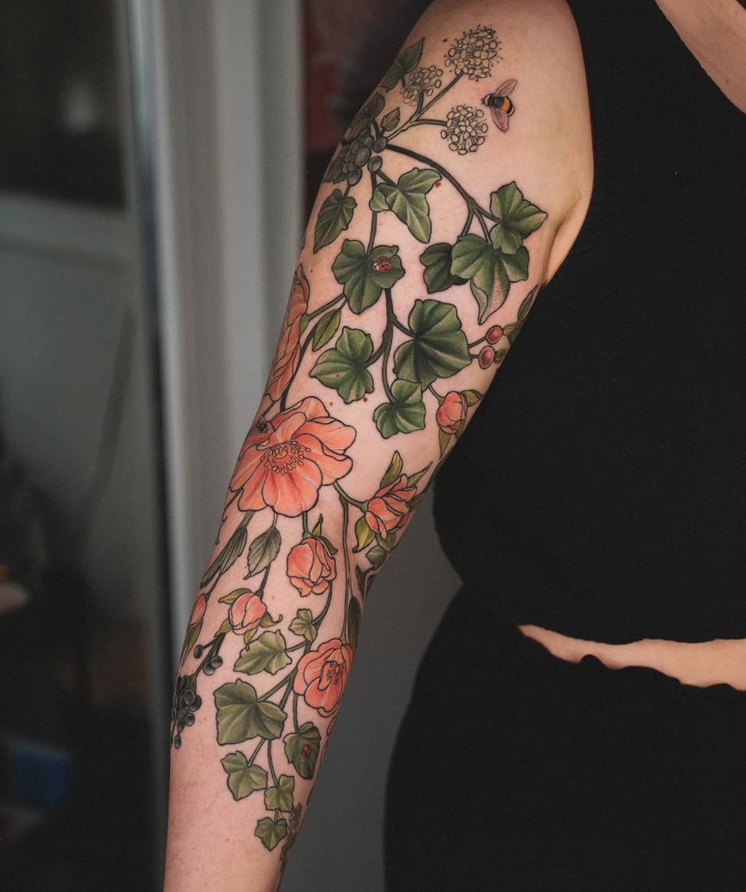 Full-sleeve arm tattoo featuring climbing vines with green leaves, coral flowers, white Queen Anne's lace clusters, and a bumblebee on pale skin.