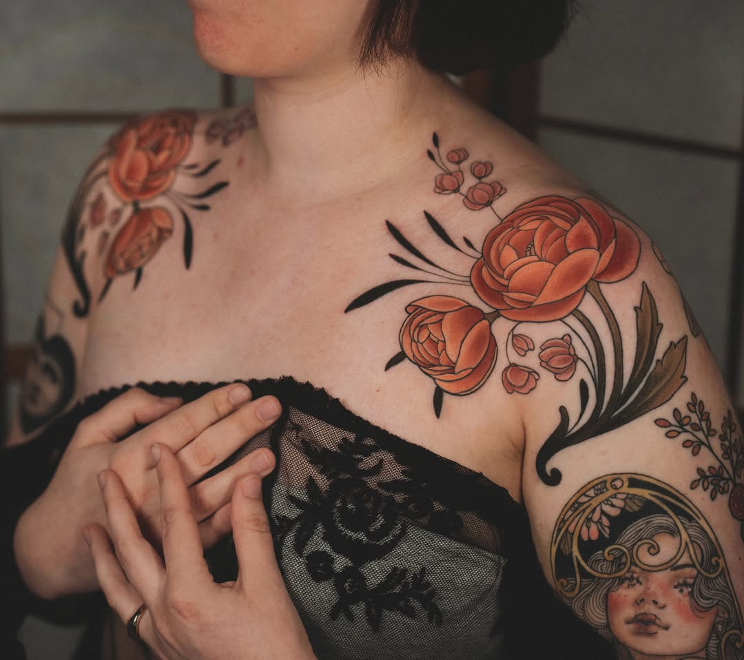 Woman's shoulder and chest displaying coral-colored roses with black stems and leaves, alongside smaller botanical elements and a portrait tattoo on the forearm.