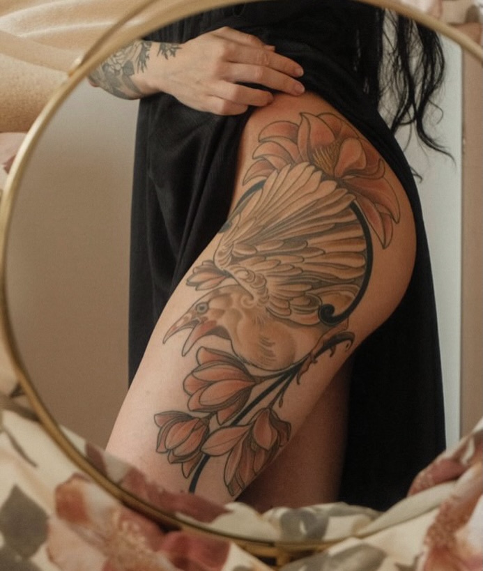 Person displaying a large thigh tattoo featuring a bird with detailed feathers and floral botanical elements in brown and tan tones, photographed in a mirror.