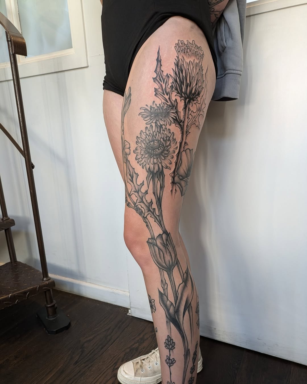 Full-leg botanical tattoo featuring detailed gerbera daisies and stems in dark ink on pale skin, photographed from the side.