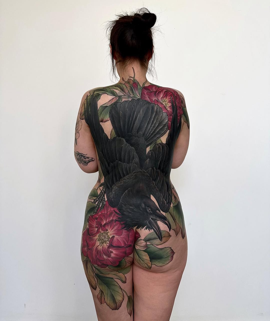 Back tattoo featuring a black raven with spread wings surrounded by large red peonies and green foliage in full color.