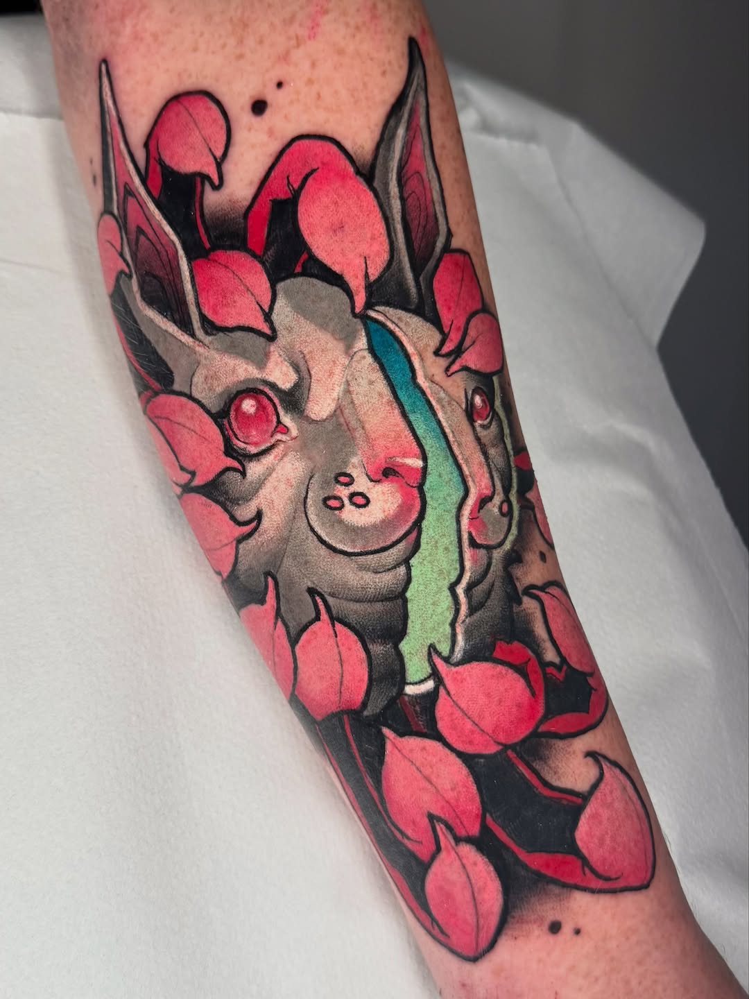 Colorful tattoo on forearm featuring a horned creature surrounded by red petals and flowers with green and tan shading details.