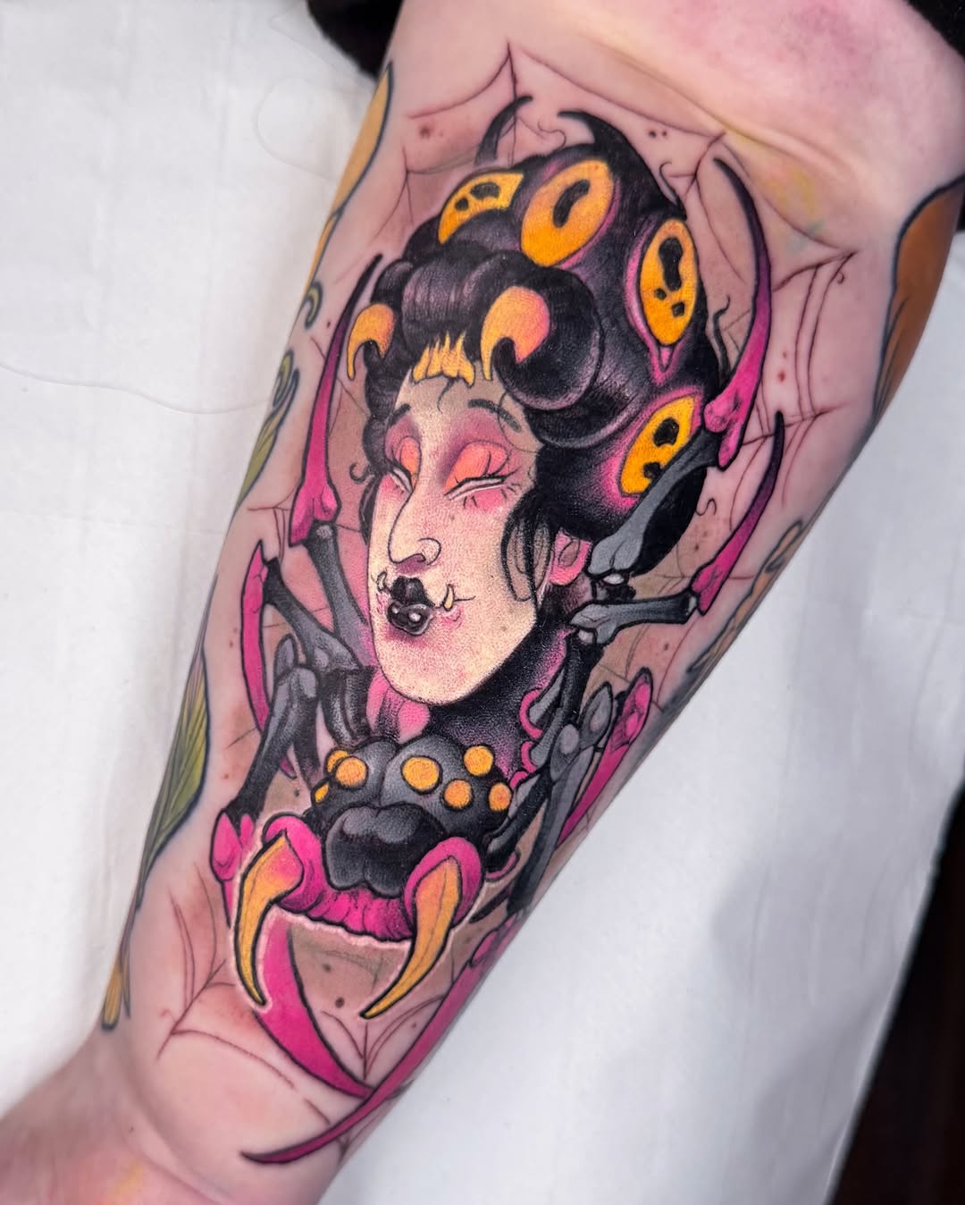 Colorful tattoo on forearm featuring a woman's face with closed eyes and black hair adorned with orange and yellow circular elements, surrounded by pink and magenta decorative shapes.