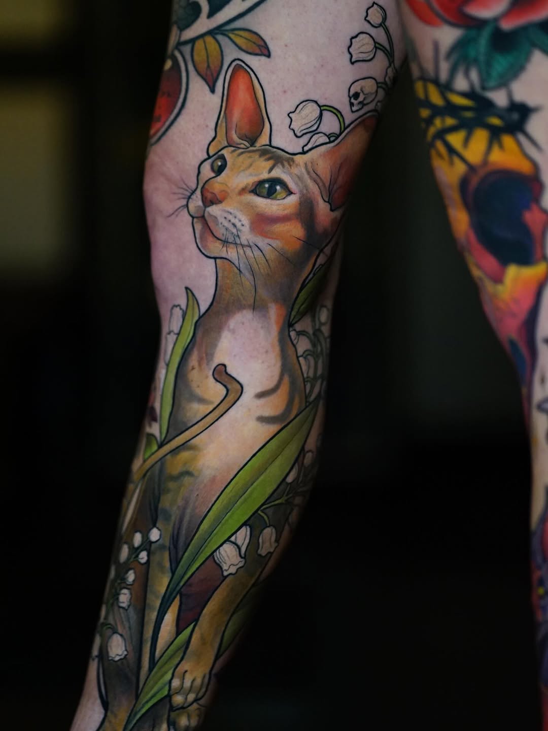 A tabby cat with green eyes surrounded by lily of the valley flowers and botanical elements tattooed on the forearm in warm brown and green tones.