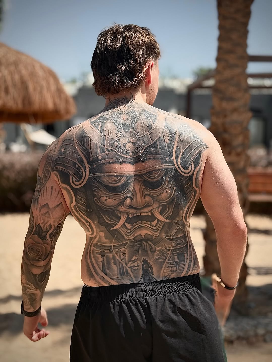 Shirtless man displaying extensive back and arm tattoos featuring a demon mask, roses, and intricate detailed designs in brown and black ink.