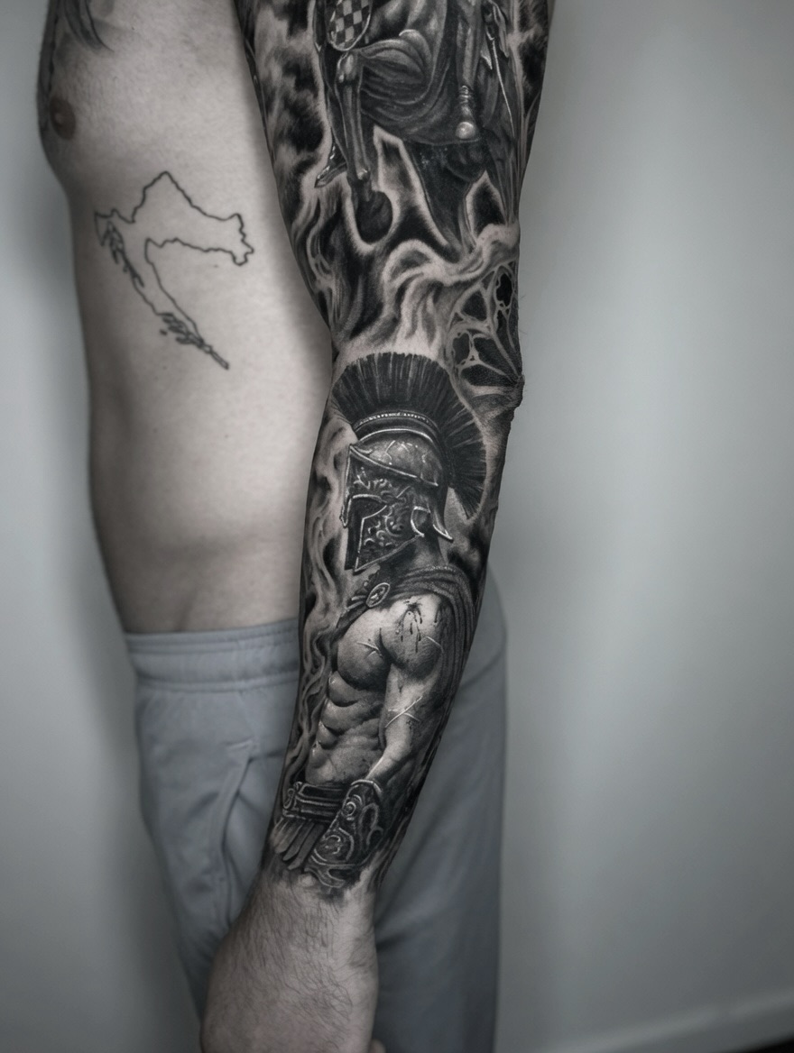 Two legs displaying a detailed sleeve tattoo featuring classical warriors, mythological figures, and ornate armor rendered in grayscale on the right leg and a simple outline map on the left.