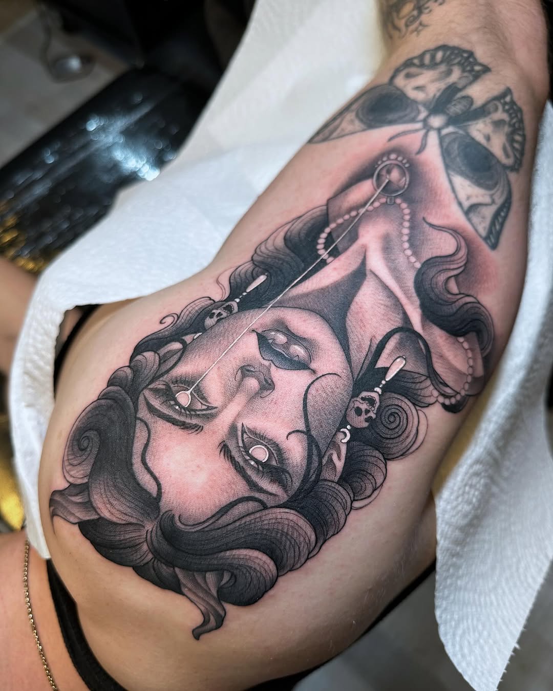 Detailed portrait tattoo on upper arm featuring a face with closed eyes, ornate jewelry, and flowing hair in black and pink shading.