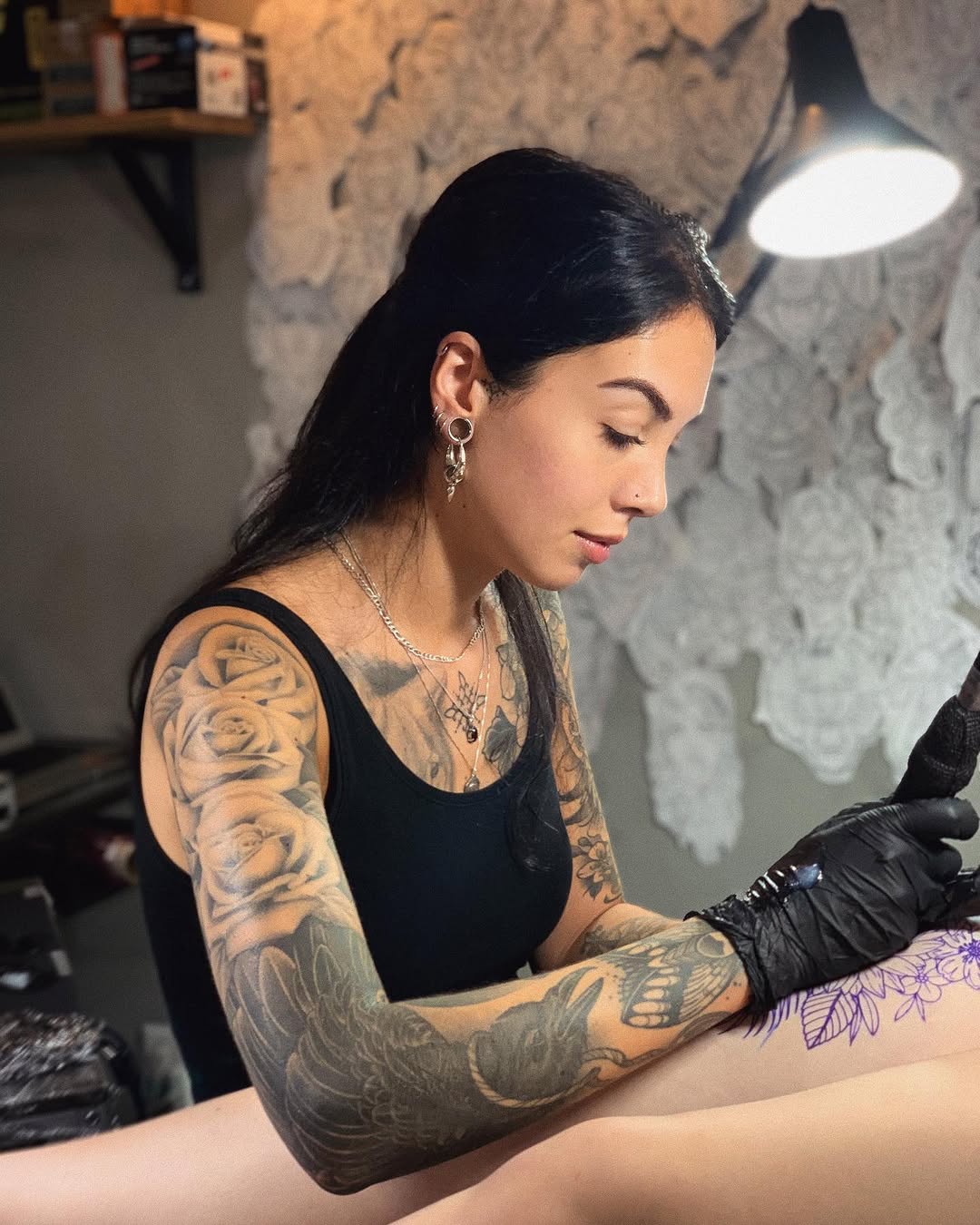 Tattoo artist applies purple floral design to client's forearm using black ink while wearing gloves under studio lighting.