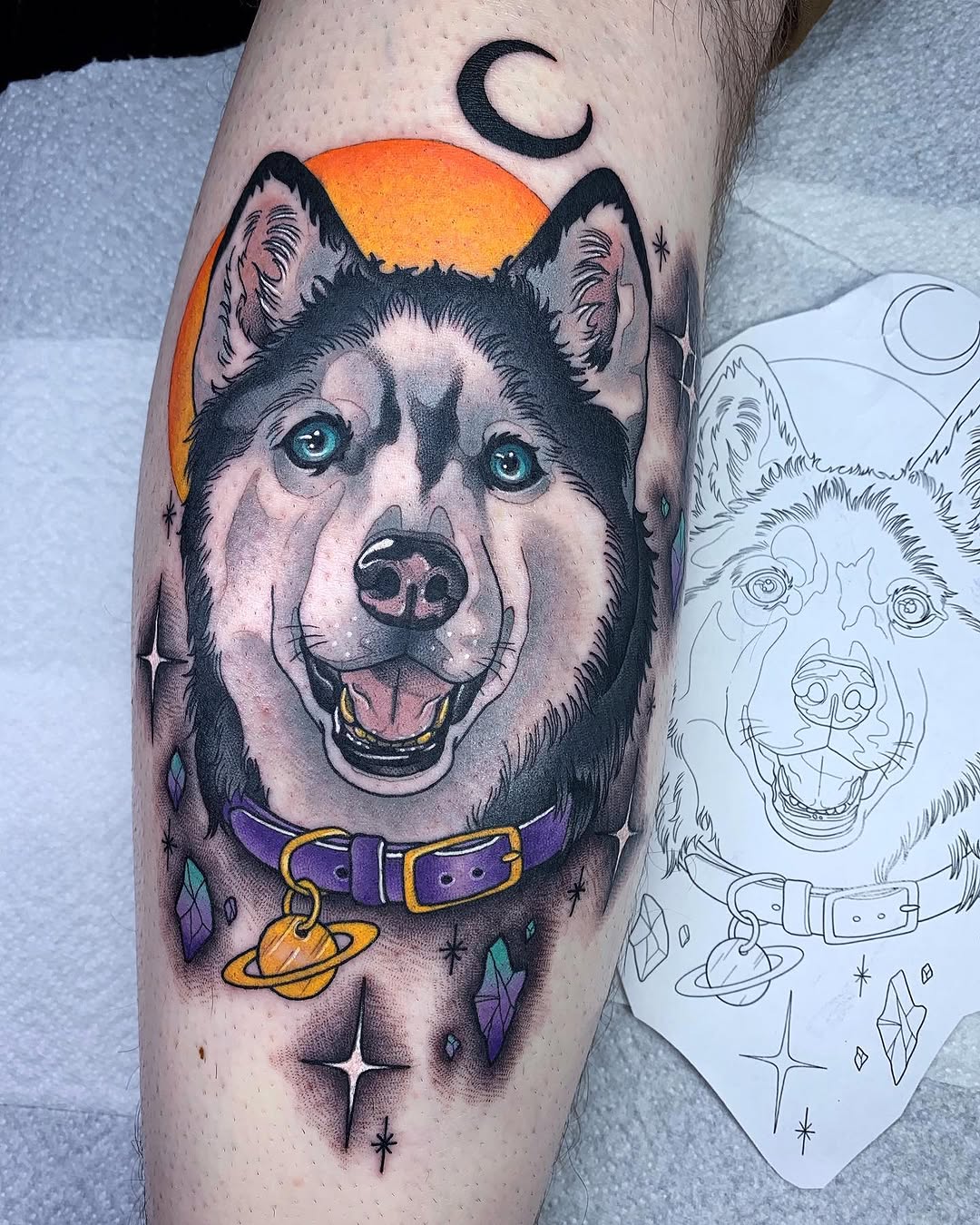 Colorful husky dog portrait tattoo on leg featuring purple collar with gold buckle, orange planet, crescent moon, and scattered stars.