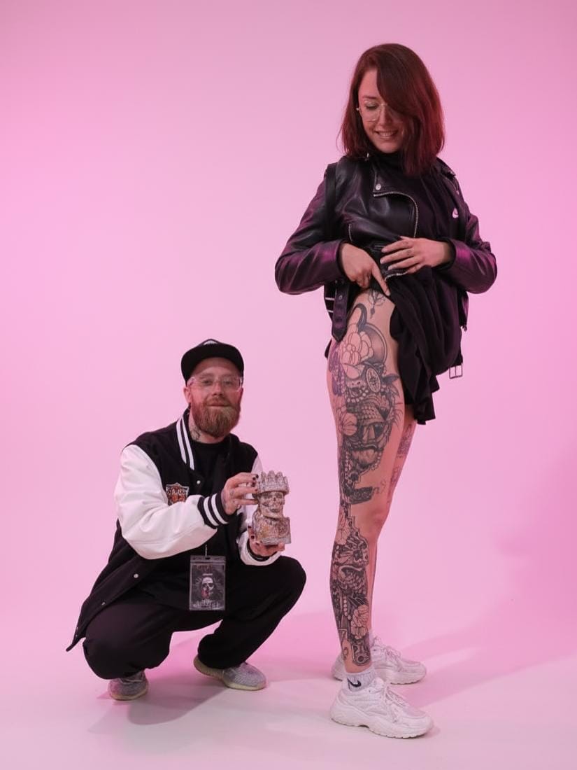 Tattoo artist displays extensive leg piece featuring ornate designs and decorative elements in dark ink on client against pink background.