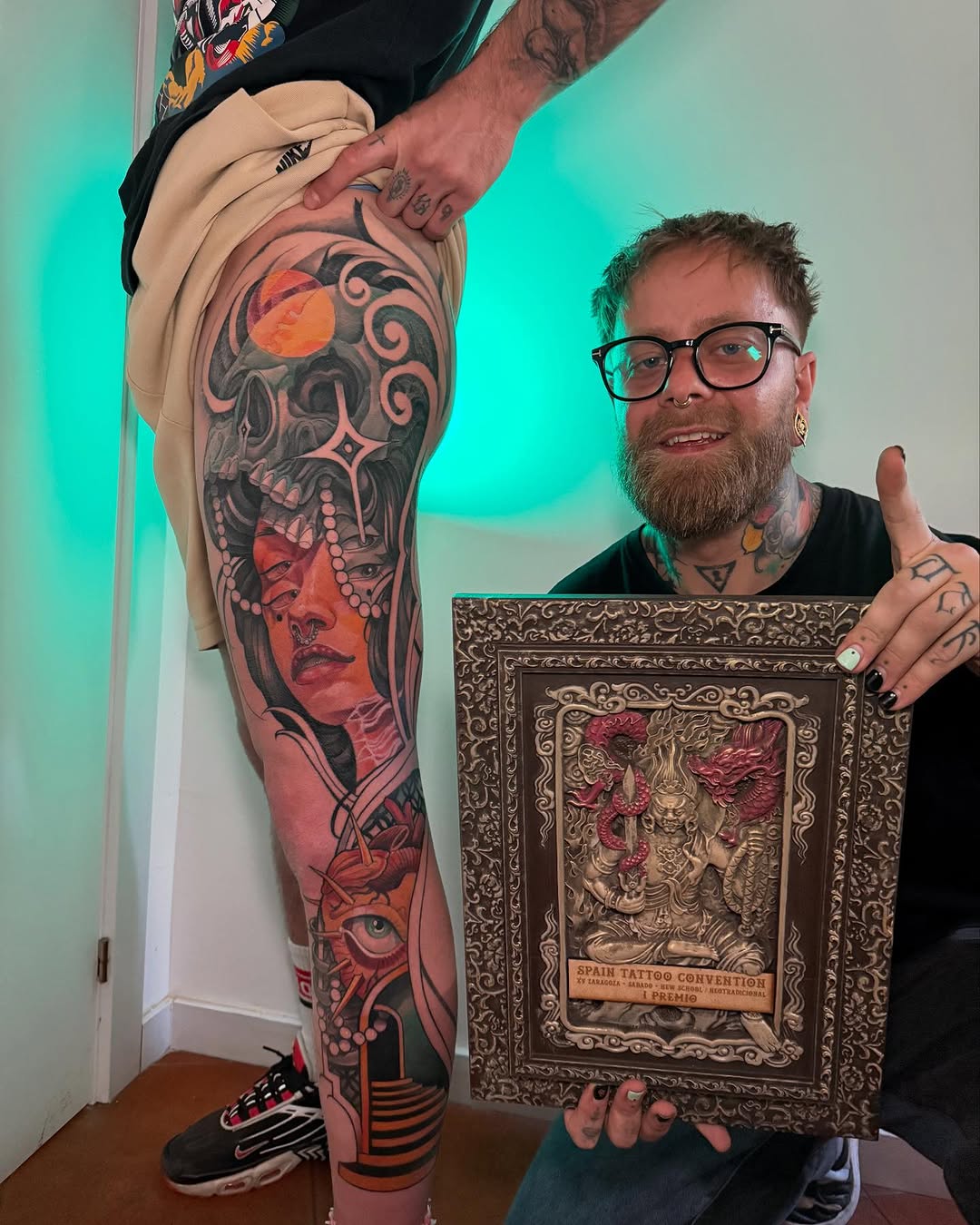 Tattoo artist displays full leg sleeve featuring a woman's face with skull imagery in red and black ink, holding a framed award.