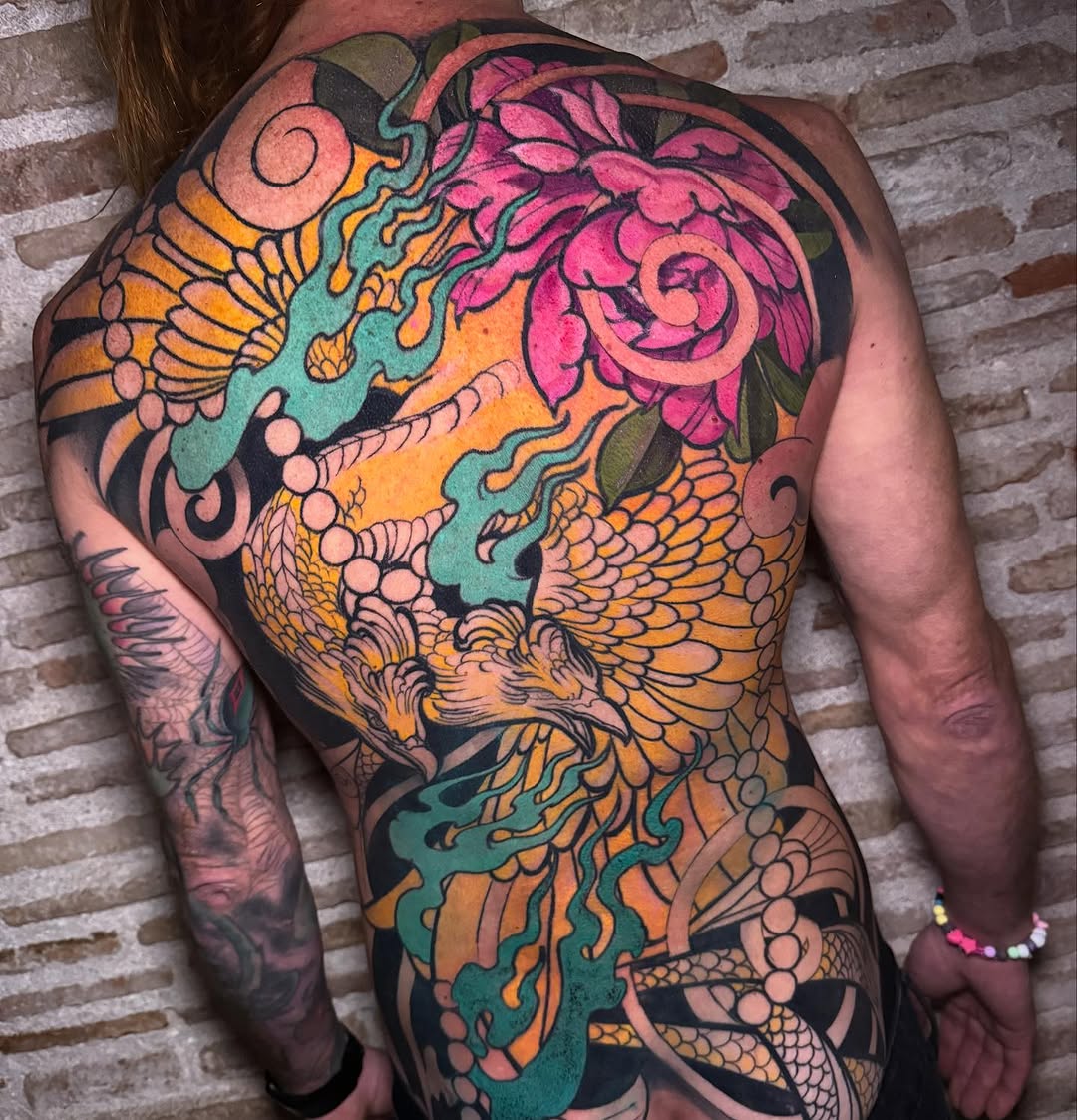 Colorful back tattoo featuring winged creatures, pink flowers, teal flames, and orange details with black outlines against brick background.