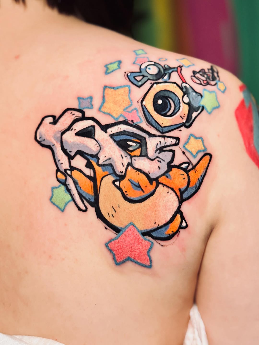Colorful tattoo on shoulder featuring a cartoon character with large eyes holding binoculars, surrounded by multicolored stars in orange, blue, yellow, and red.