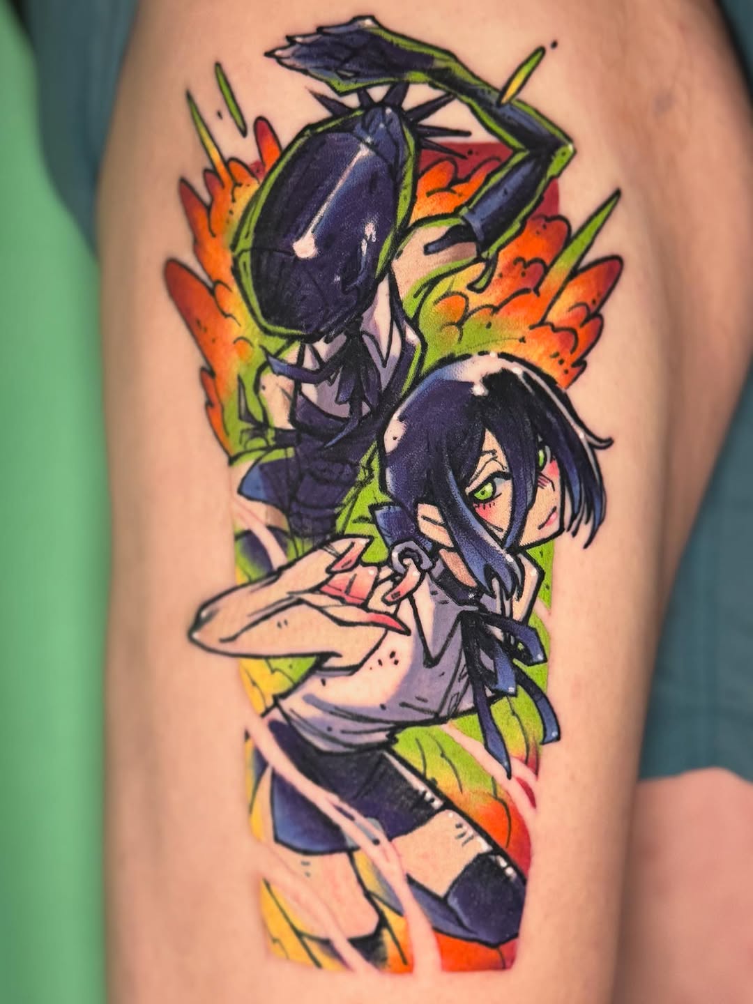 Colorful upper arm tattoo featuring an anime character with dark hair and a black eggplant above, surrounded by vibrant orange, green, and yellow flames.